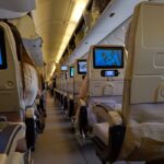 View of an airplane cabin interior with passengers seated, featuring screens on seatbacks.